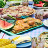 Tabletop summer food of burgers, watermelon, pizza, with drinks and people eating outside.