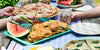 Tabletop summer food of burgers, watermelon, pizza, with drinks and people eating outside.